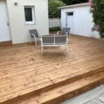 photo d'une grande terrasse avec deux marches, collée à une maison et habillée d'un salon de jardin compsoé de deux chaises et une double assise gris et blanc.